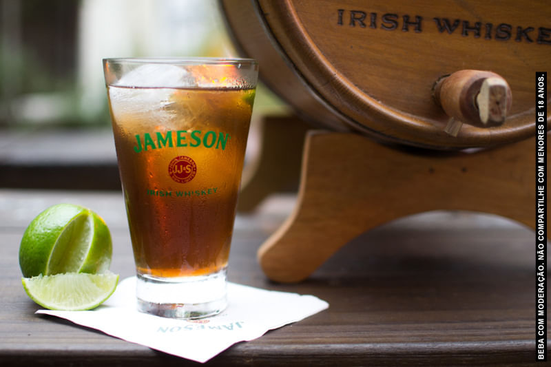 Um copo de Jameson com drink preparado com chá aparece em destaque em cima da mesa, junto com um limão fatiado e um barril de whiskey